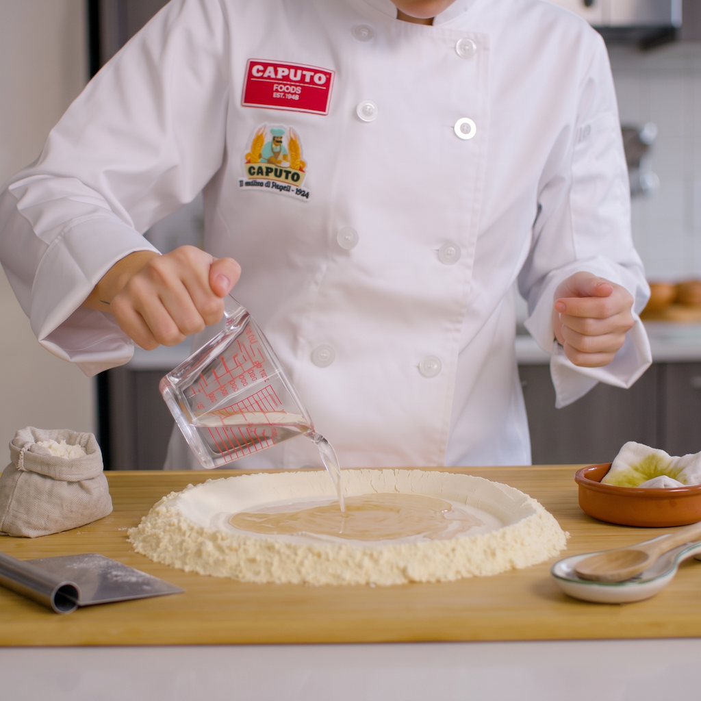 A chef in a white uniform pours liquid from a glass measuring cup onto flour spread in a shallow circle on a wooden board, with baking tools nearby and a CAPUTO logo patch visible on the chest.