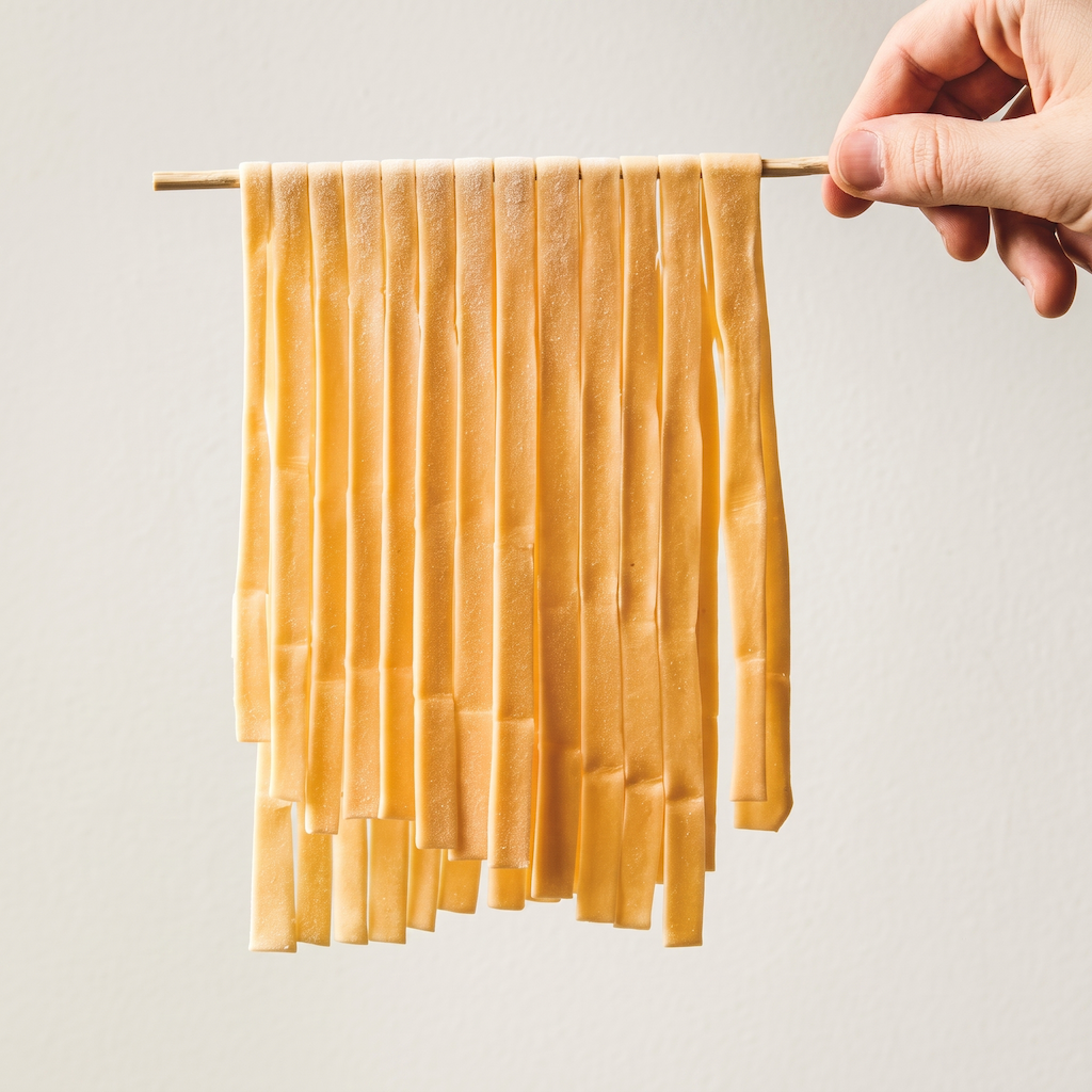 A hand holding a thin wooden rod with multiple narrow strips of pale yellow fabric or ribbon hanging from it, shown against a plain light gray background.