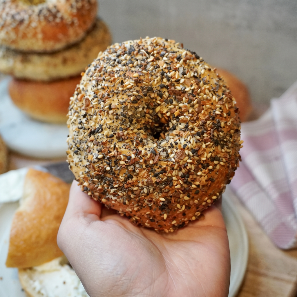 A hand holding a seeded bagel in the foreground, with more bagels and a slice of bagel and cream cheese on a table in the background.