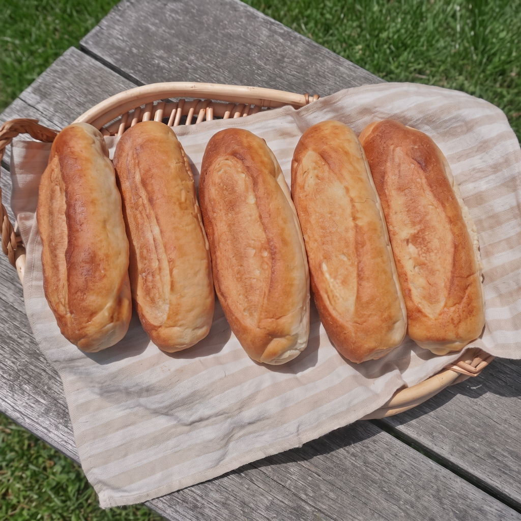 Five golden-brown baguette-style loaves of bread arranged in a wicker basket lined with a beige striped cloth, placed on a weathered gray wooden table outdoors with green grass visible in the background.