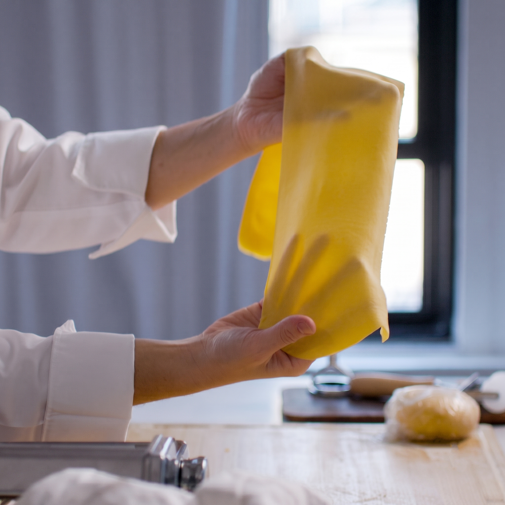 A person in a white chef's coat holds up a sheet of yellow pasta dough over a kitchen counter with baking tools in the background.