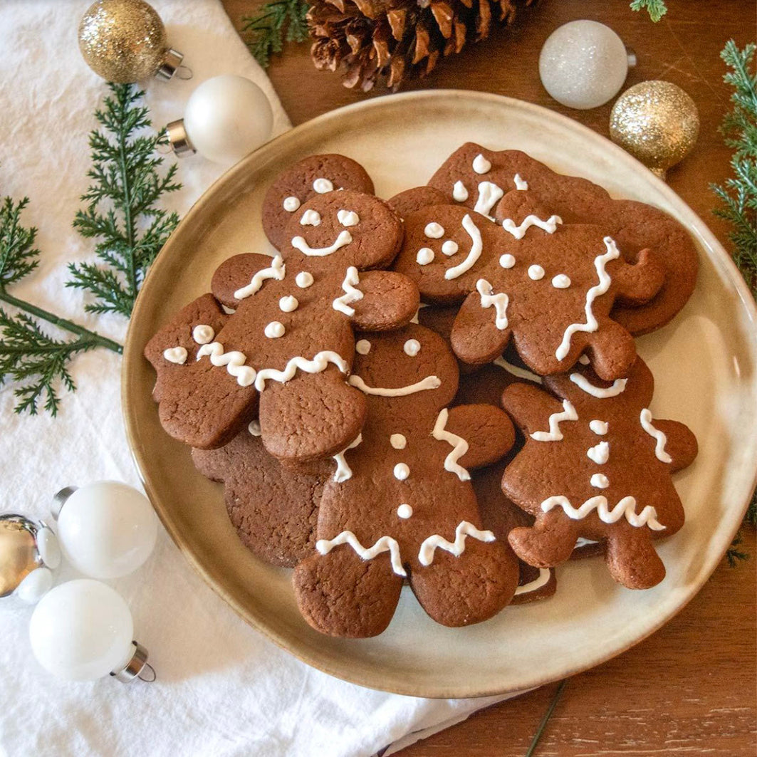 A plate of decorated gingerbread cookies shaped like gingerbread men and trees, arranged on a wooden table with holiday ornaments, pinecones, and greenery.