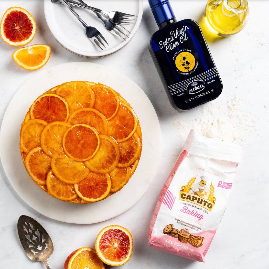 A styled flat-lay featuring a round orange-topped cake on a white plate, a bottle of extra virgin olive oil, a bag of Caputo Baking flour, sliced blood oranges, and utensils on a white surface.