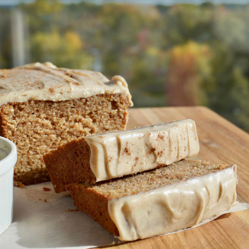 A loaf of moist pumpkin spice cake with a thick layer of light beige frosting, shown with two slices on a wooden board. A small white ramekin is partially visible on the left, and a blurred outdoor background with trees is in focus behind the cake.