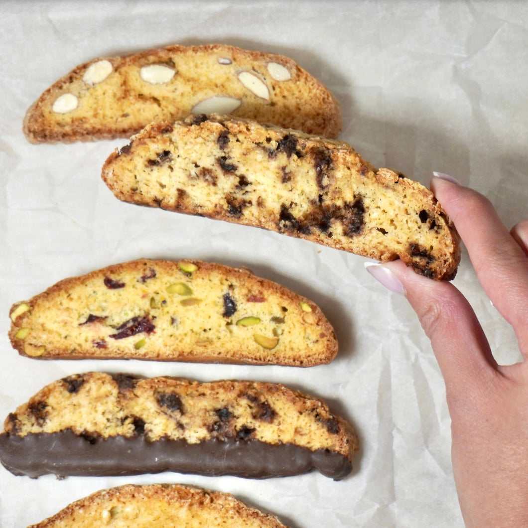 Five almond biscotti cookies of various flavors and toppings are arranged on parchment paper, with a hand holding one cookie to show its texture.