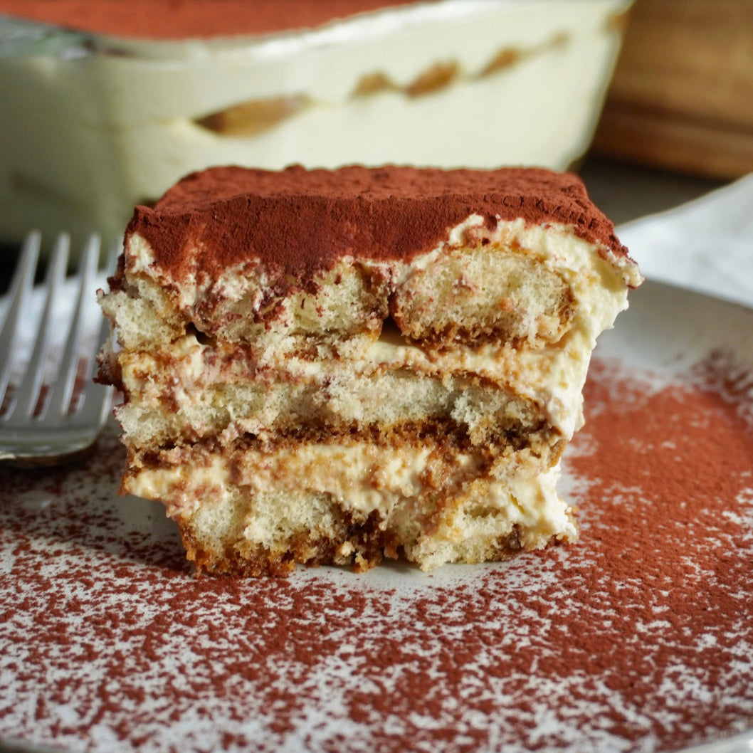 A close-up of a slice of tiramisu dessert on a speckled plate, with layers of cream and sponge cake visible and cocoa powder dusting the top and surrounding surface.