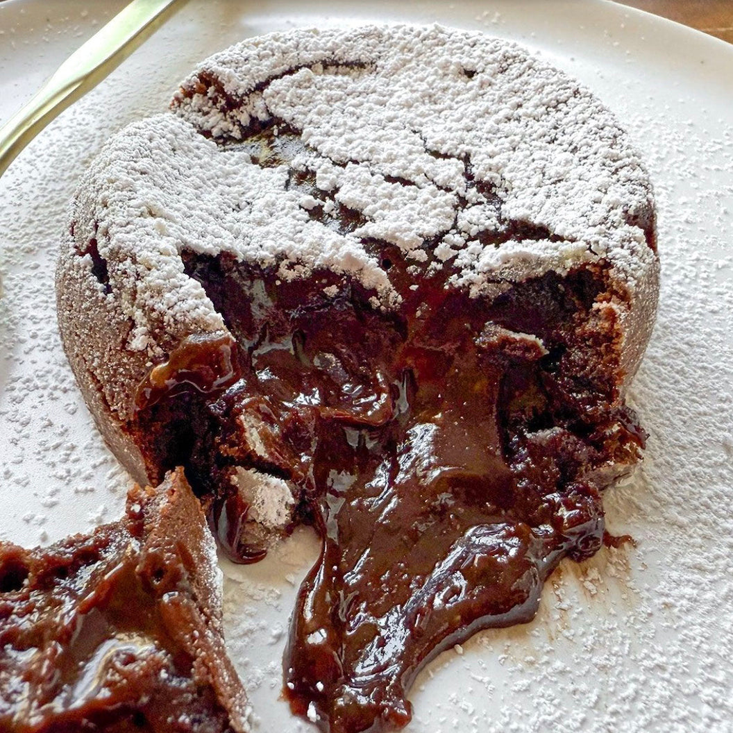 A close-up of a molten chocolate lava cake on a white plate, dusted with powdered sugar and with a piece broken off to show the glossy, dripping chocolate center.