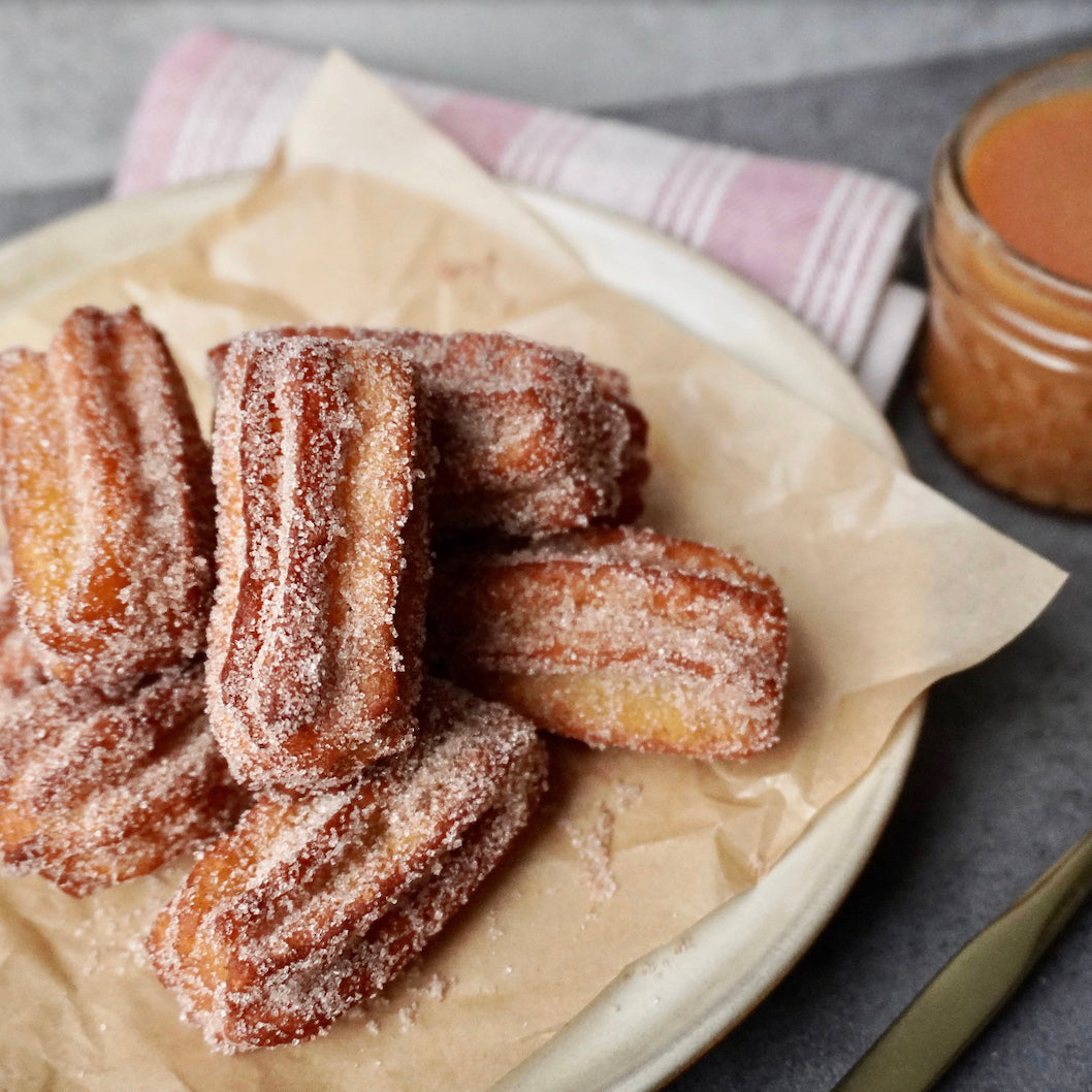 A plate of golden-brown fried dough sticks dusted with sugar, served on parchment paper with a striped napkin and a small jar of sauce in the background.