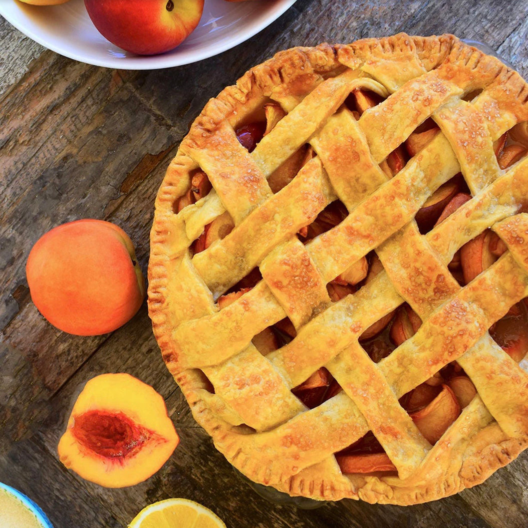 A golden-brown lattice-crust pie filled with fruit sits on a rustic wooden table, surrounded by fresh peaches, a sliced peach, and a lemon wedge. A white plate with more fruit is visible in the upper left.