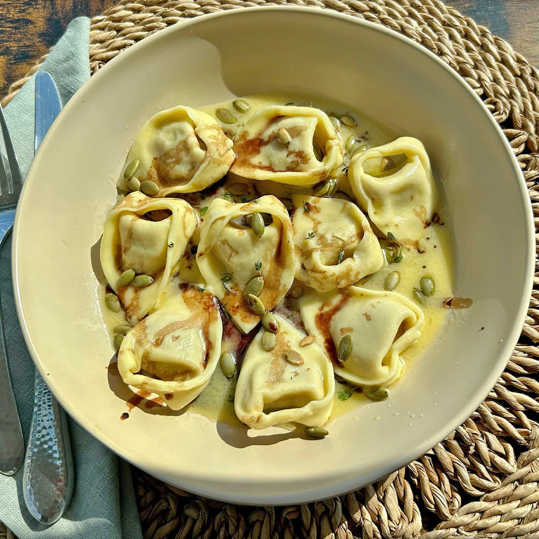 A bowl of stuffed pasta tortellini in a creamy yellow sauce, topped with pumpkin seeds, served on a woven placemat with a knife and napkin visible.