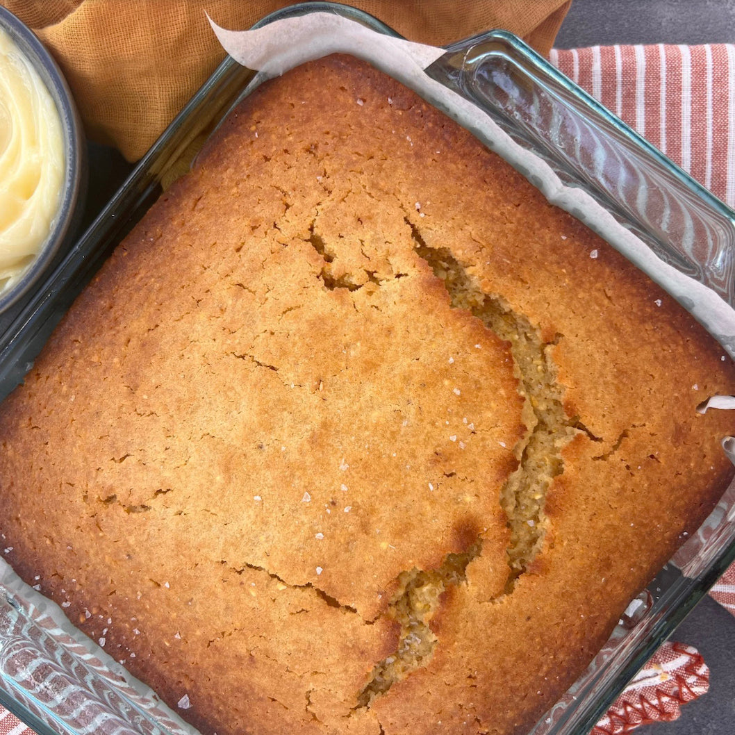 A freshly baked square loaf of golden-brown bread in a clear glass baking dish with parchment paper, shown from a top-down angle with a small bowl of butter partially visible at the upper left.