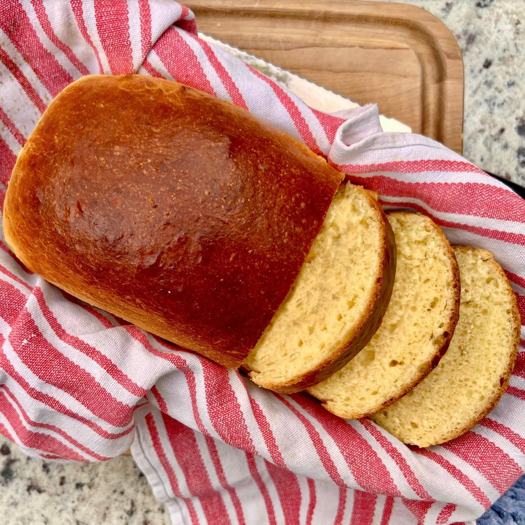 A loaf of golden-brown bread with three slices cut, resting on a red and white striped kitchen towel on a wooden cutting board over a speckled countertop.