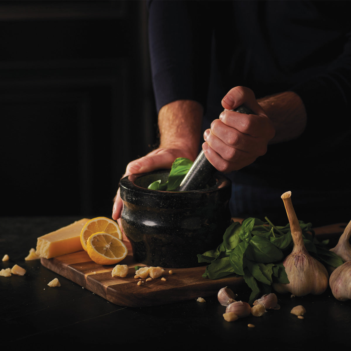 Person using a mortar and pestle with lemons, garlic, and herbs on a dark surface