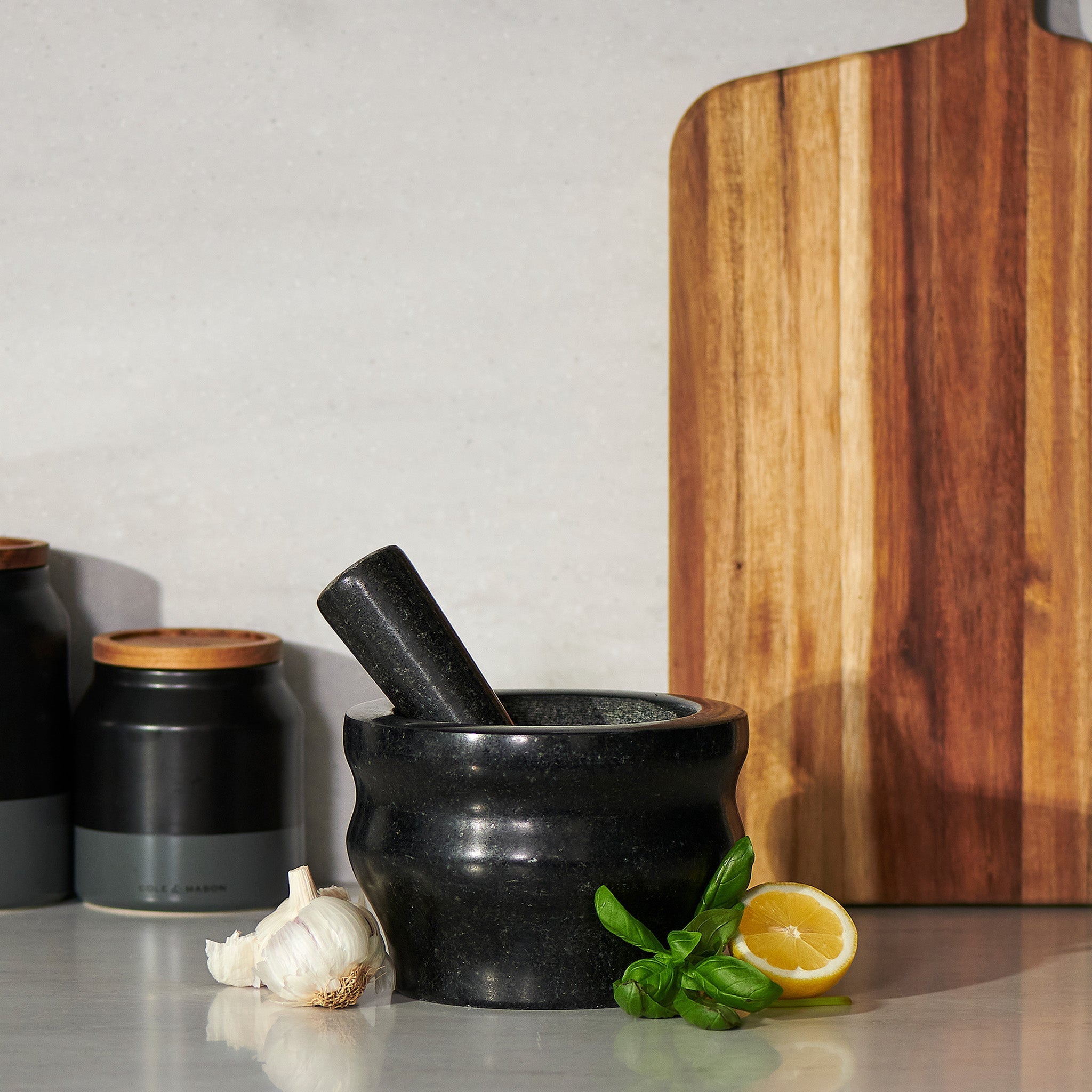 Black mortar and pestle on a kitchen counter with a wooden cutting board and ingredients.