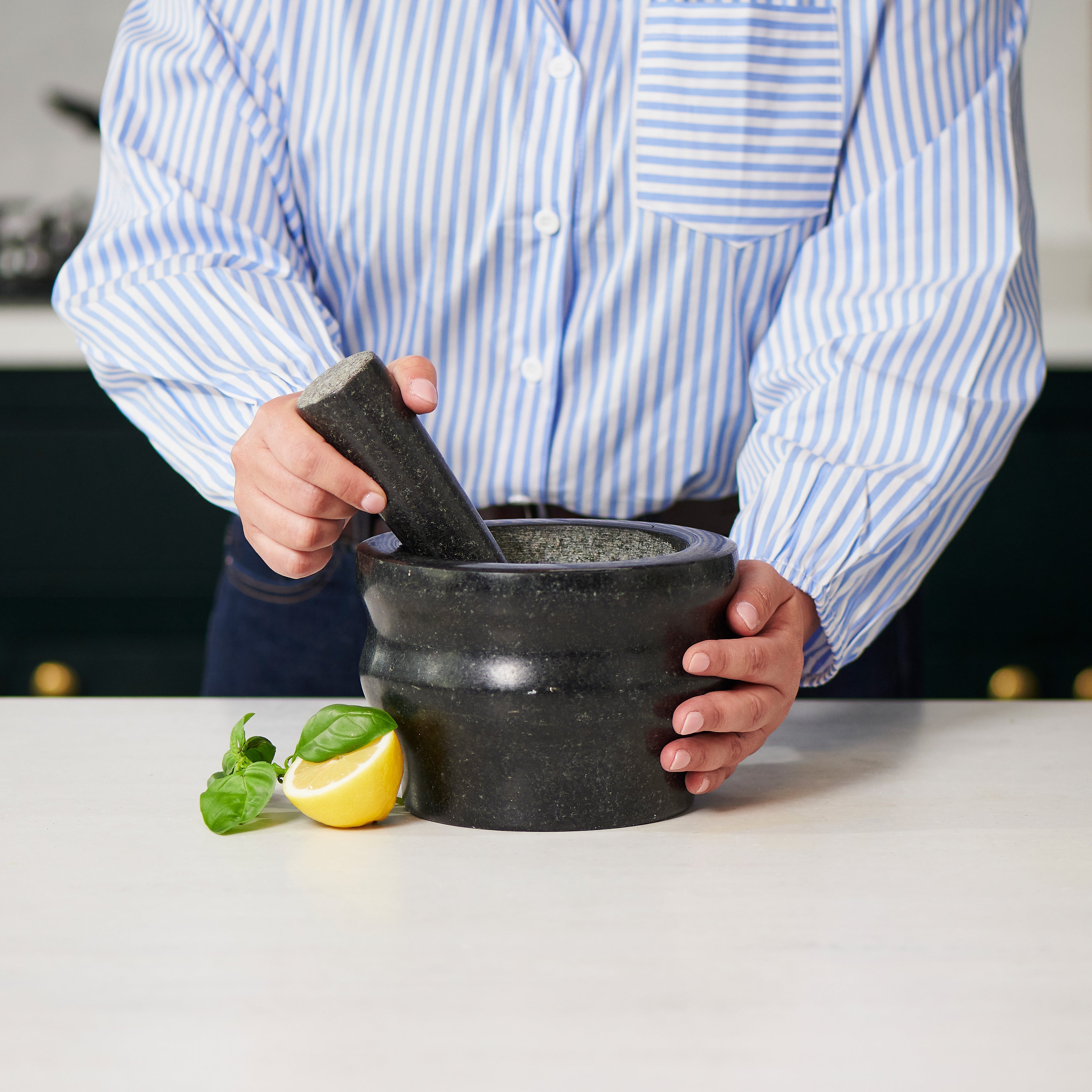 Person using a mortar and pestle on a kitchen counter with a lemon and herbs in the background.