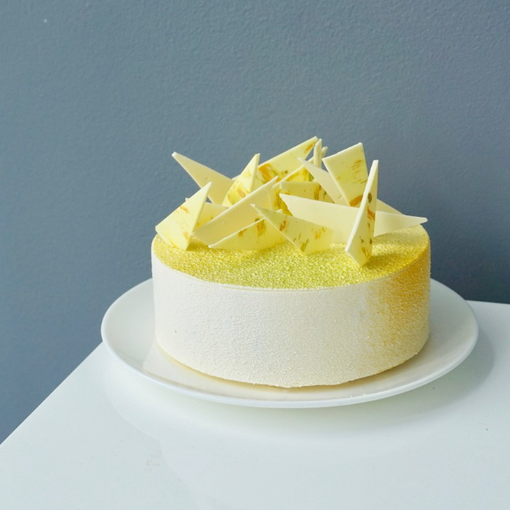 A round frosted cake on a white plate, decorated with pale yellow durian powder and white chocolate shards on top, set against a dark blue-gray wall and white surface.