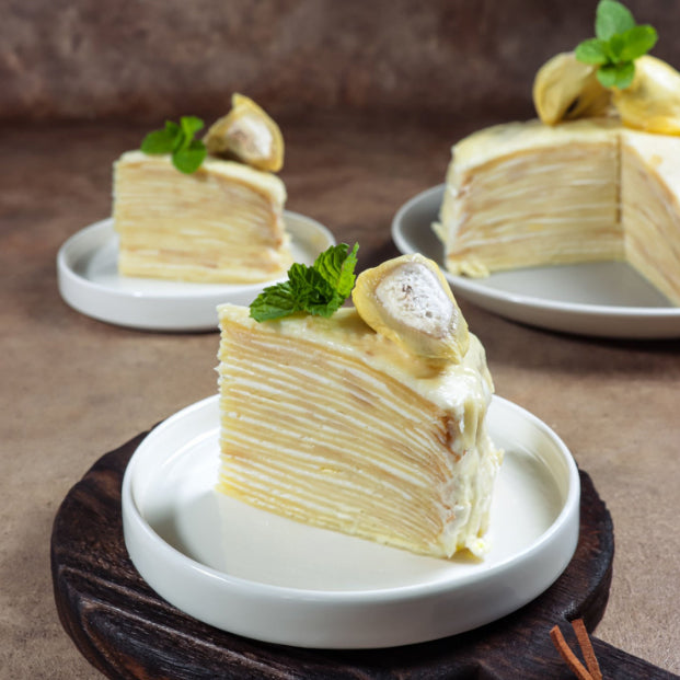 Three slices of layered crepe cake with durian powder and mint garnish, served on white plates against a warm brown background.