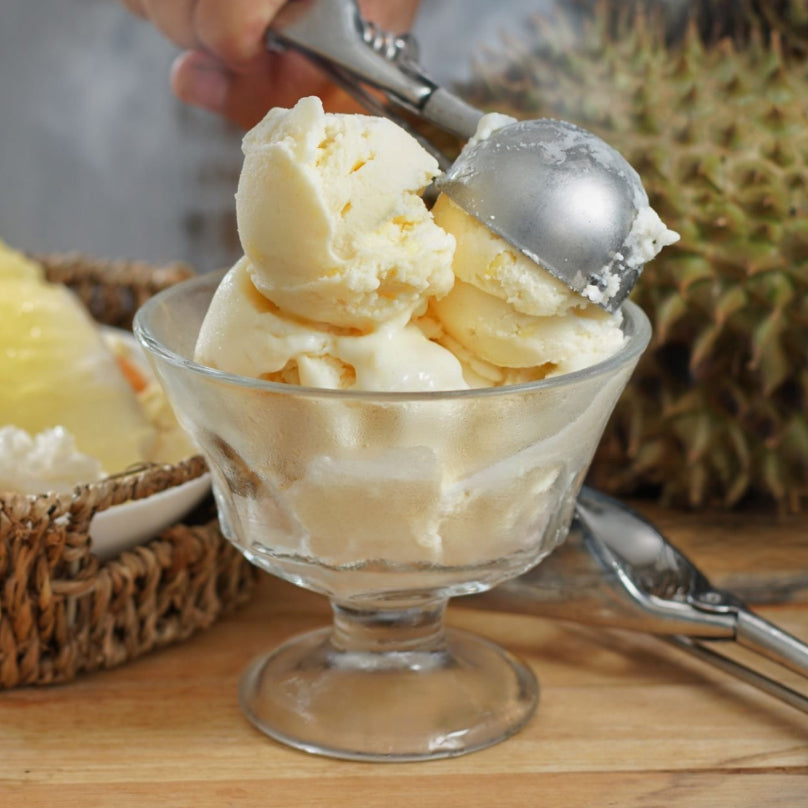 Scoops of creamy durian ice cream with durian powder garnish in a clear glass sundae bowl on a wooden surface, with a metal ice cream scoop being held above the bowl and a pineapple in the background.