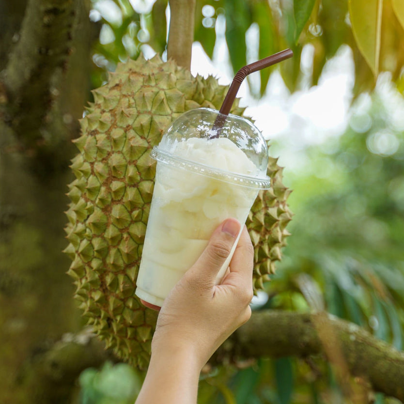 A hand holds a clear plastic cup of durian drink garnished with durian freeze dried powder, with a dome lid and bent brown straw in front of a large durian fruit hanging on a tree branch outdoors.