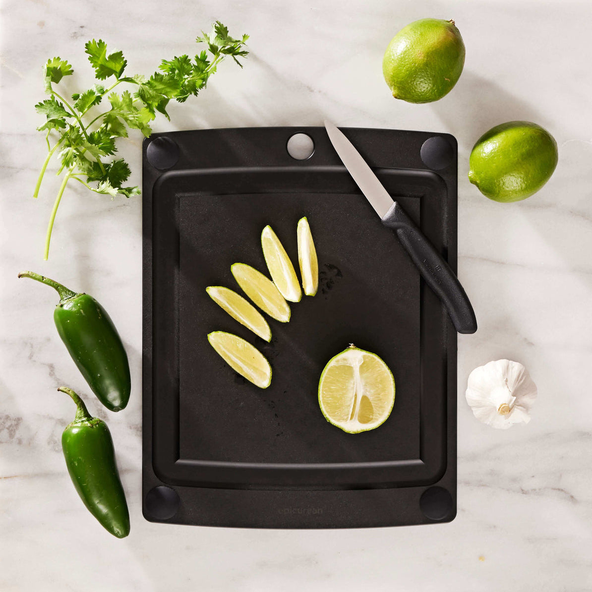 Black Epicurean cutting board with non-slip feet with sliced lemons, a knife, and garnishes on a marble surface