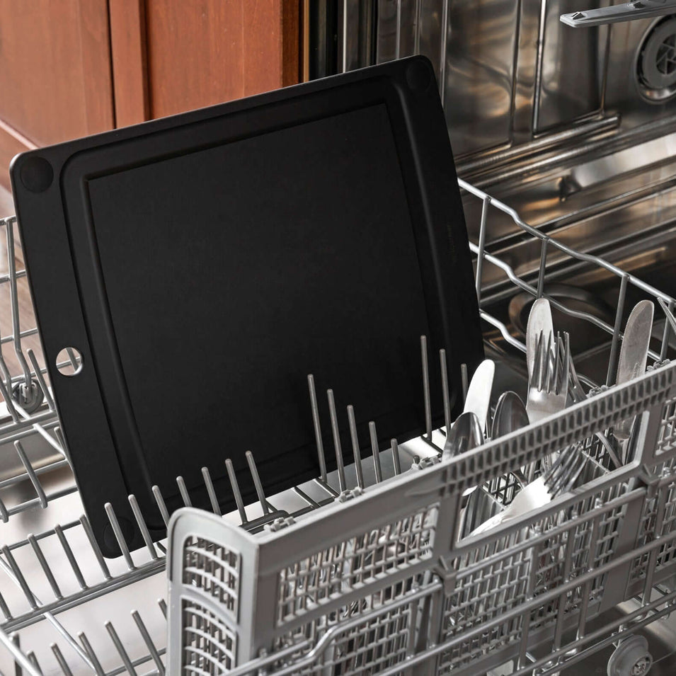 Black Epicurean non-slip all-in-one series cutting board inside an open dishwasher with cutlery and plates.