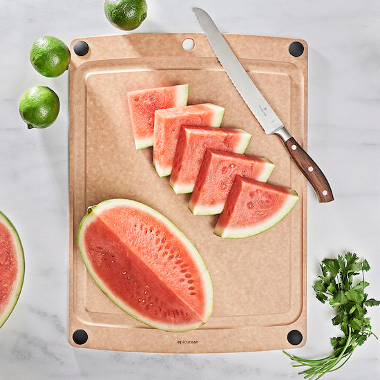 Sliced watermelon on a Epicurean non-slip all-in-one series cutting board with a knife, limes, and herbs on a marble surface.