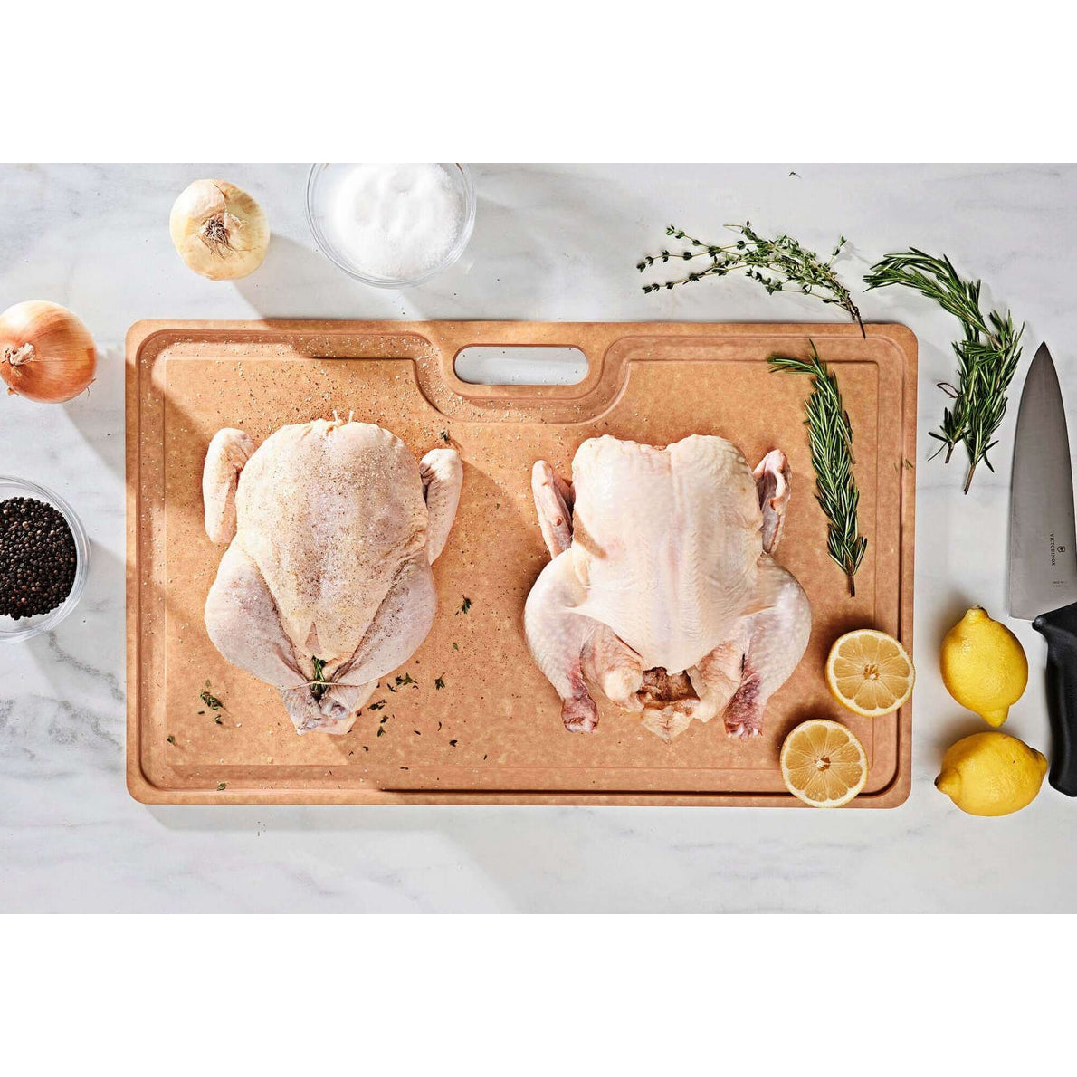 Two raw chickens on a wooden Epicurean cutting board with herbs and lemons on a white marble surface.
