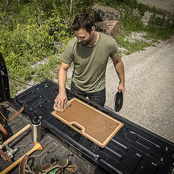 Man preparing food on a Epicurean butcher cutting board in the back of a pickup truck with nature in the background