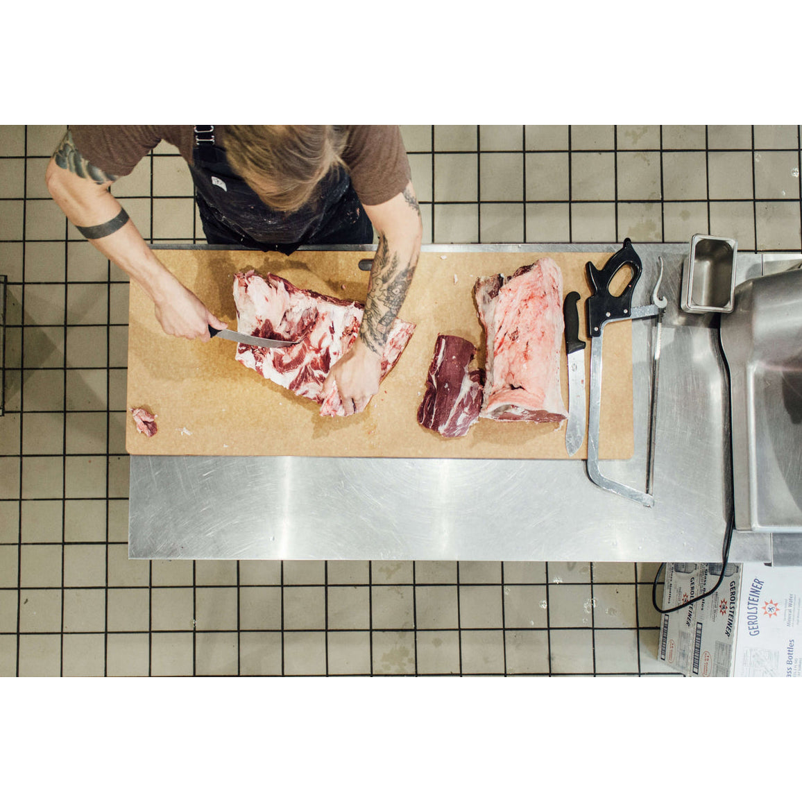 Person cutting raw meat on a wooden Epicurean big game and butcher cutting board with various tools in a kitchen setting.