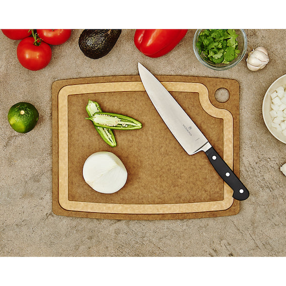Epicurean nutmeg cutting board with a knife, vegetables, and herbs on a stone surface