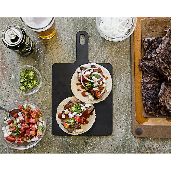 Two tacos on a black Epicurean Handy series cutting board with a beer and ingredients on a rustic surface.