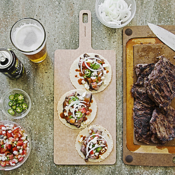 Tortillas with various toppings on a Epicurean Handy wooden board, accompanied by a glass of beer and a cutting board with grilled meat.