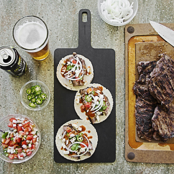 Tacos on a black Epicurean Handy cutting board with a glass of beer, condiments, and a cutting board with meat on a rustic surface.