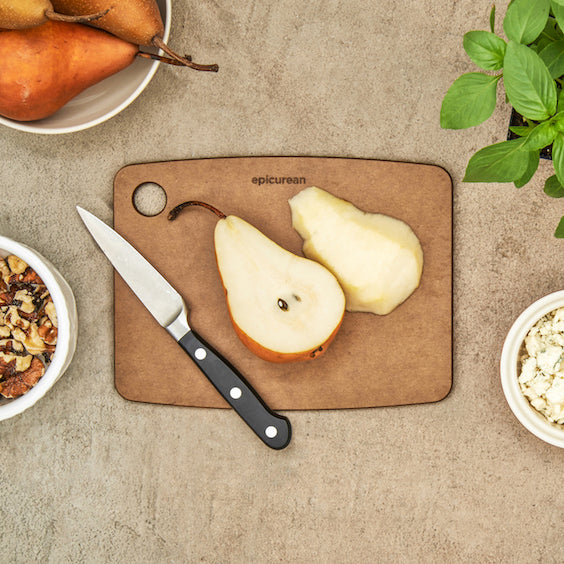 Sliced pears on a small, Nutmeg Epicurean kitchen series original cutting board with a knife, on a textured surface with bowls and a plant.