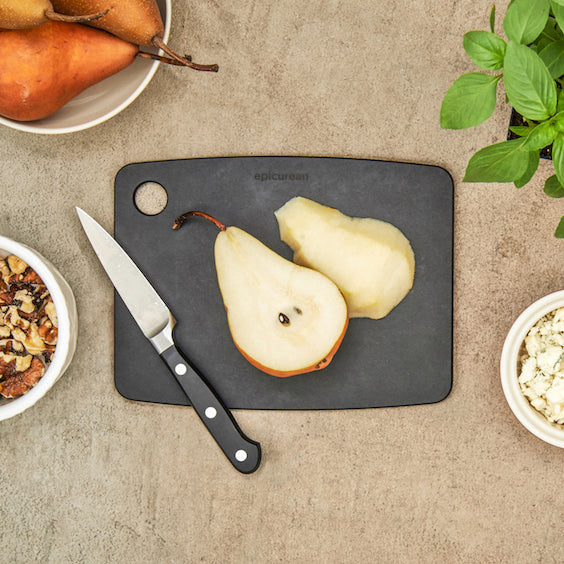 Two pears on a small, dark Epicurean kitchen series cutting board with a knife, surrounded by bowls of food and a plant.