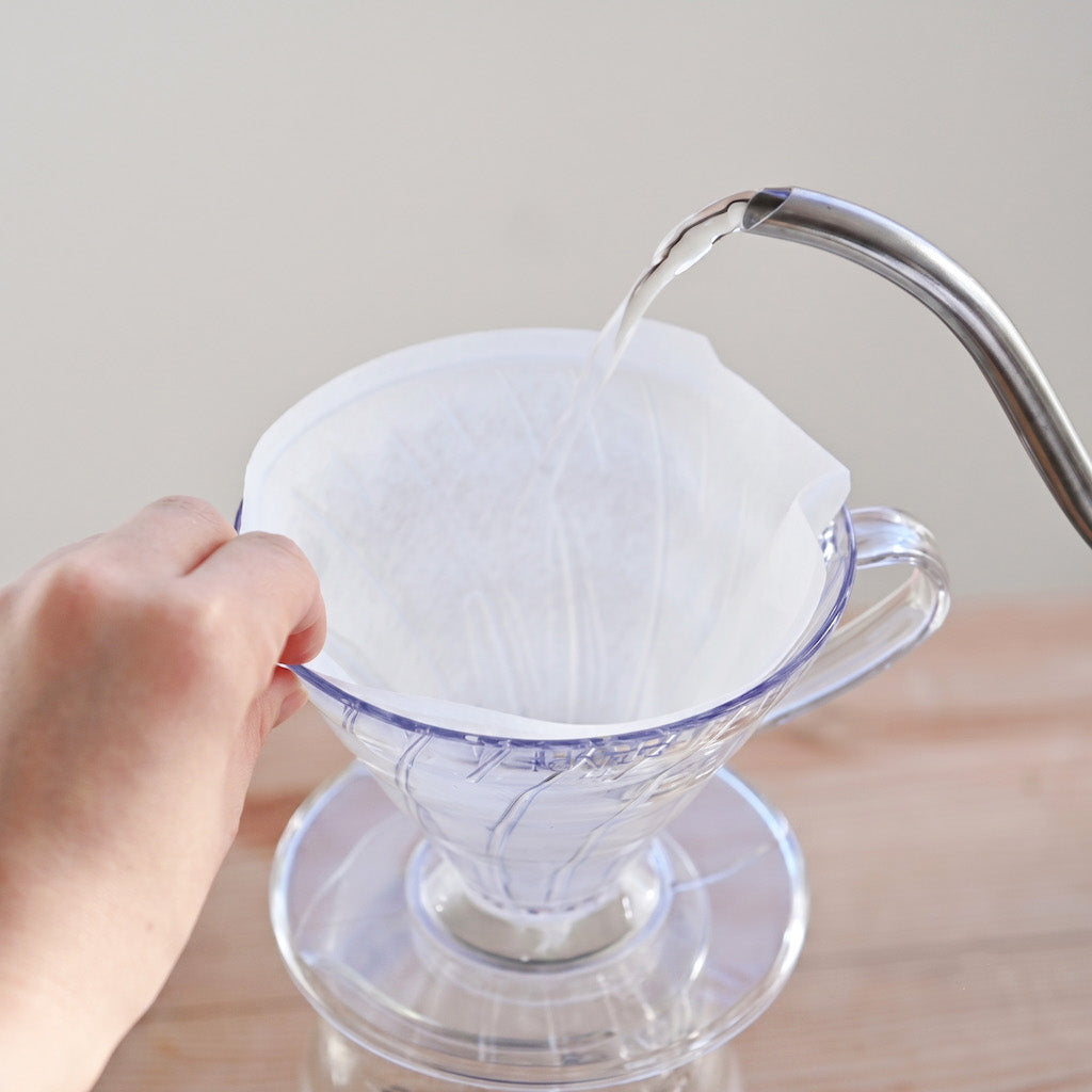 A hand holds a white paper filter cone inside a clear glass pour-over coffee dripper, with water being poured from a metal gooseneck kettle on top, set on a light wooden surface against a neutral beige background.