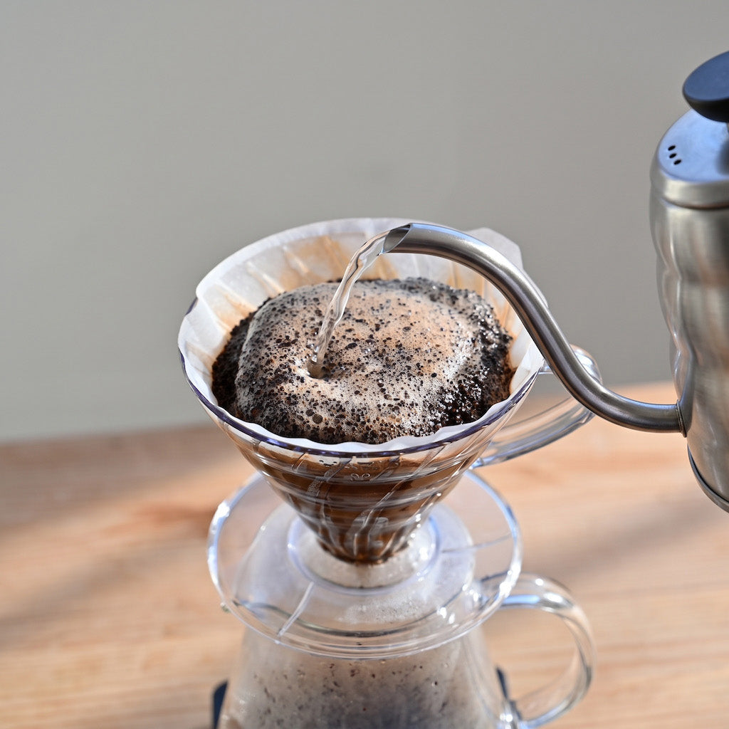 Pour-over coffee preparation with hot water being poured from a stainless steel kettle into a glass cone filter containing ground coffee, with the brewed coffee collected in a glass carafe on a wooden surface.