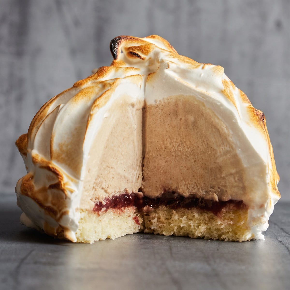 A close-up of a meringue-topped chestnut dessert cut in half, showing a light brown cream or mousse filling, a dark berry jam layer, and a biscuit or cake base on a gray surface.