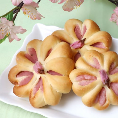 Flower-shaped bread rolls with pink Japanese sakura bean paste filling on a white plate with cherry blossoms in the background.