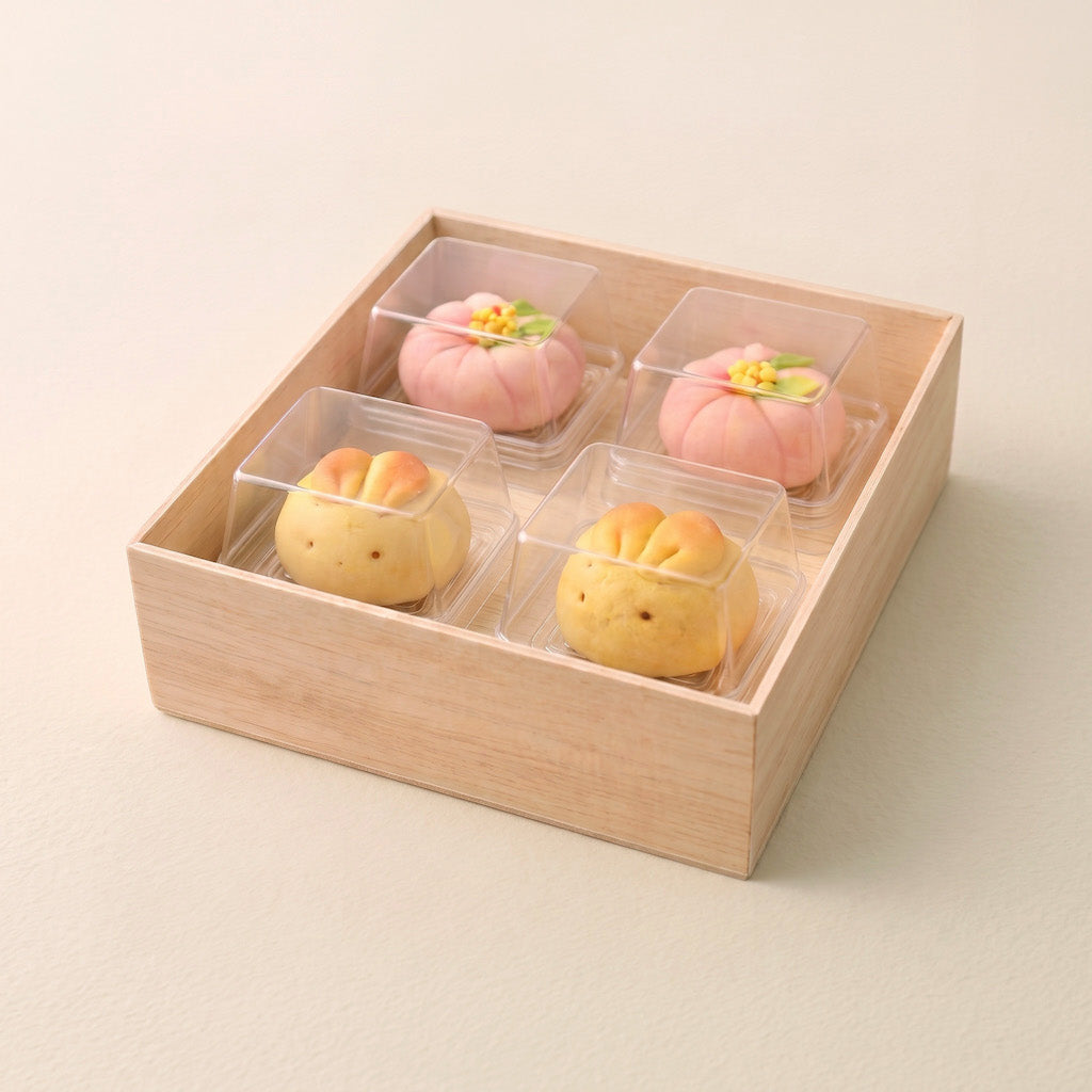 Four small decorative sweets in clear plastic containers arranged inside a light natural wood box on a plain warm beige background.