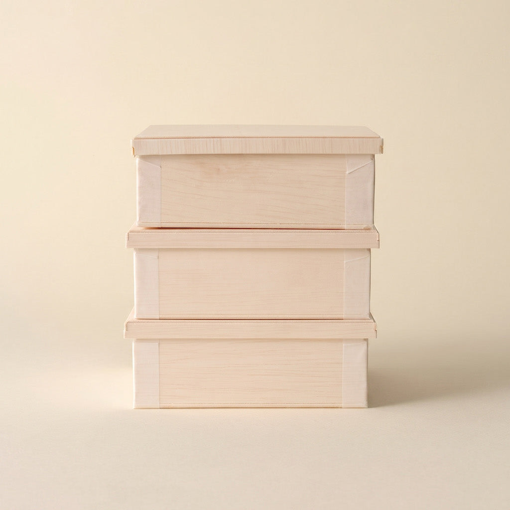 Three stacked light wood storage boxes with matching lids, set against a solid warm beige background.
