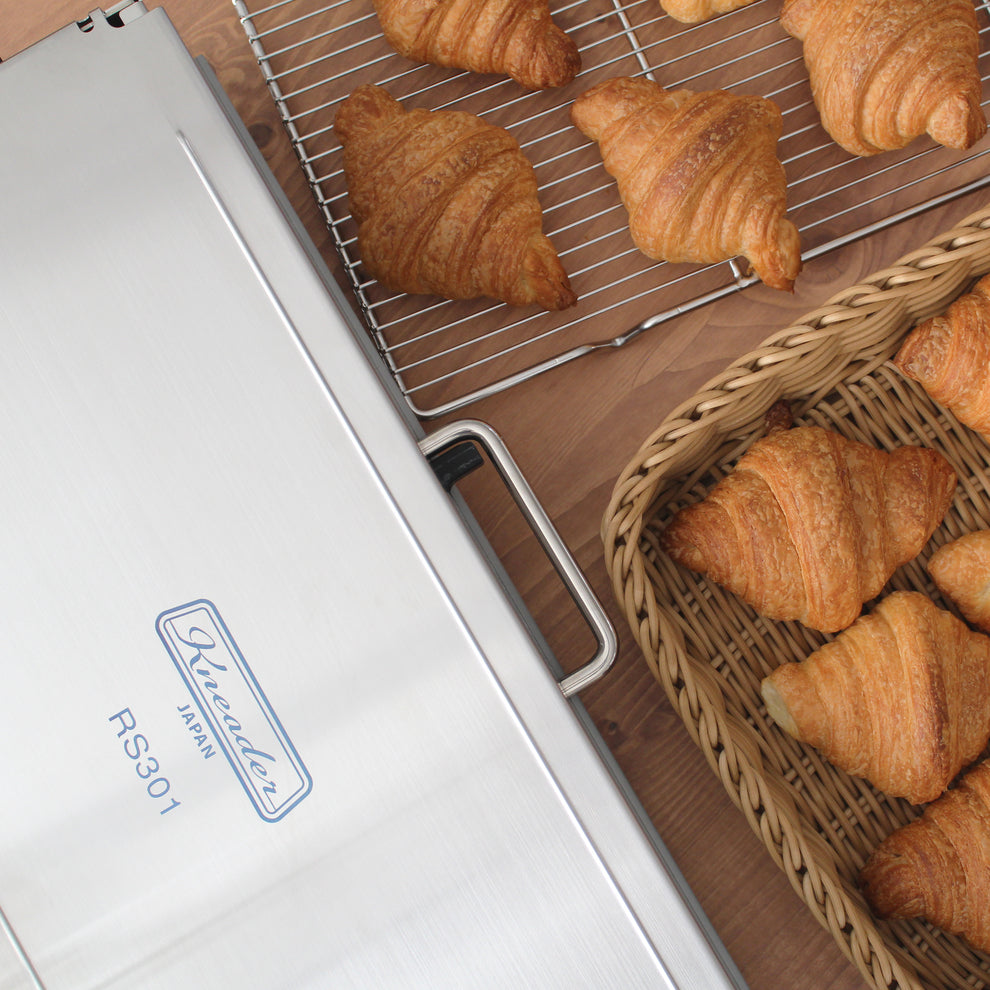 Croissants on a cooling rack and in a basket next to a Kneader sheeter holder with 'Kneader' branding.