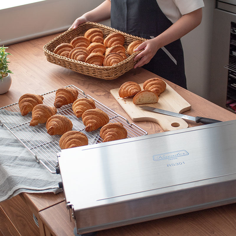 Person holding a basket of croissants next to a Kneader dough sheeter on a wooden table.