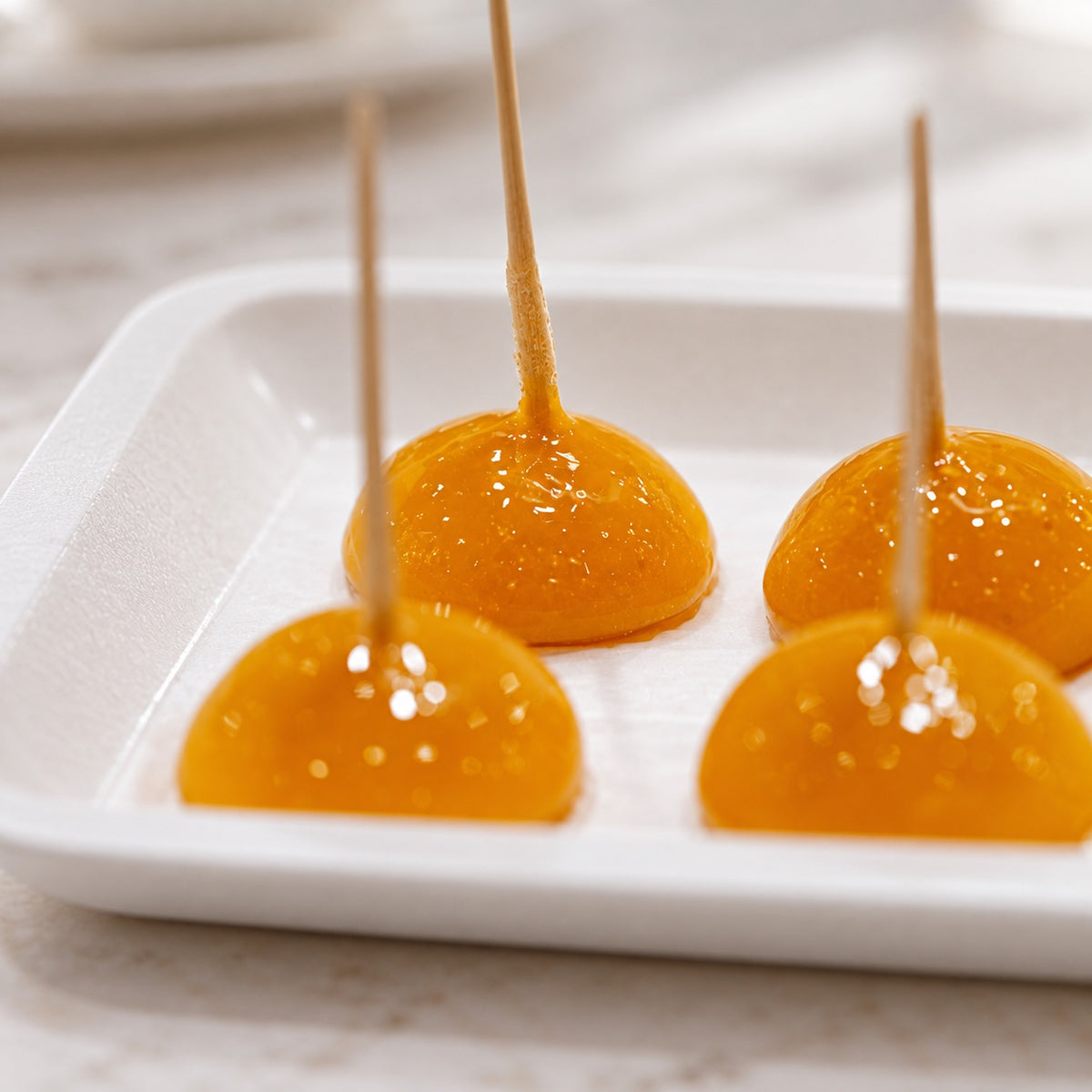 Four glossy orange gels on wooden sticks, arranged in a white square dish on a light marble surface.