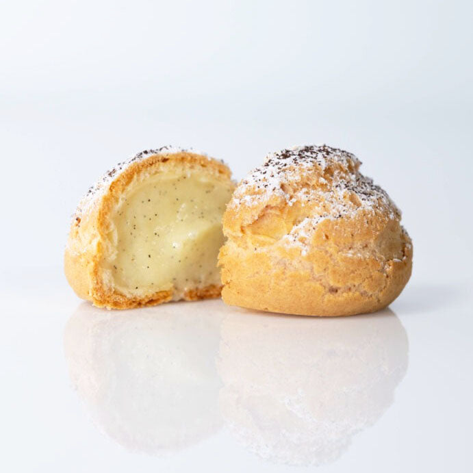 Two golden-brown cream puffs on a white reflective surface, with one cut open to show a pale custard filling and both topped with a light dusting of powdered sugar.