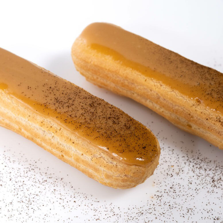 Two golden-brown choux pastry sticks topped with glossy caramel sauce and sprinkled with dark cocoa powder, set on a white surface with scattered cocoa powder.