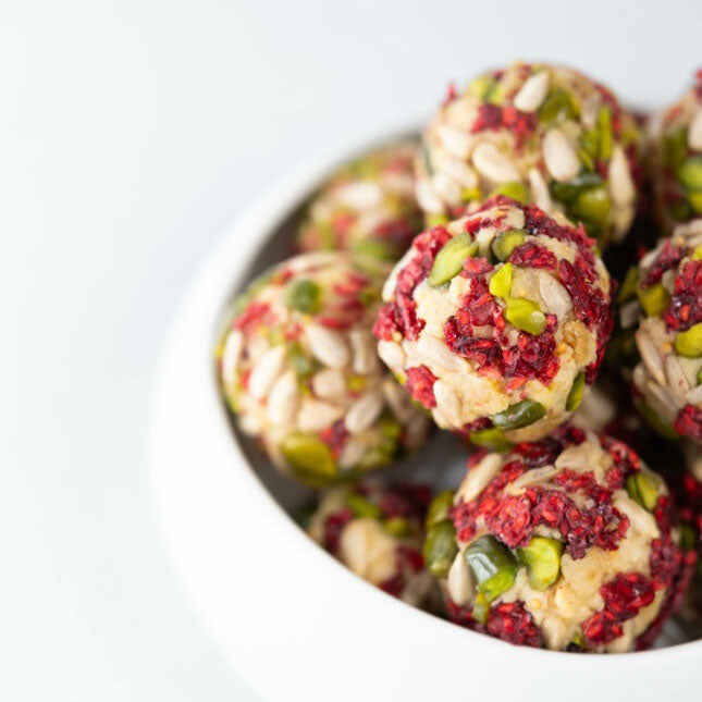 A close-up of several round desserts in a white bowl, featuring a beige base mixed with raspberry pieces and green pistachio pieces, on a clean white background.