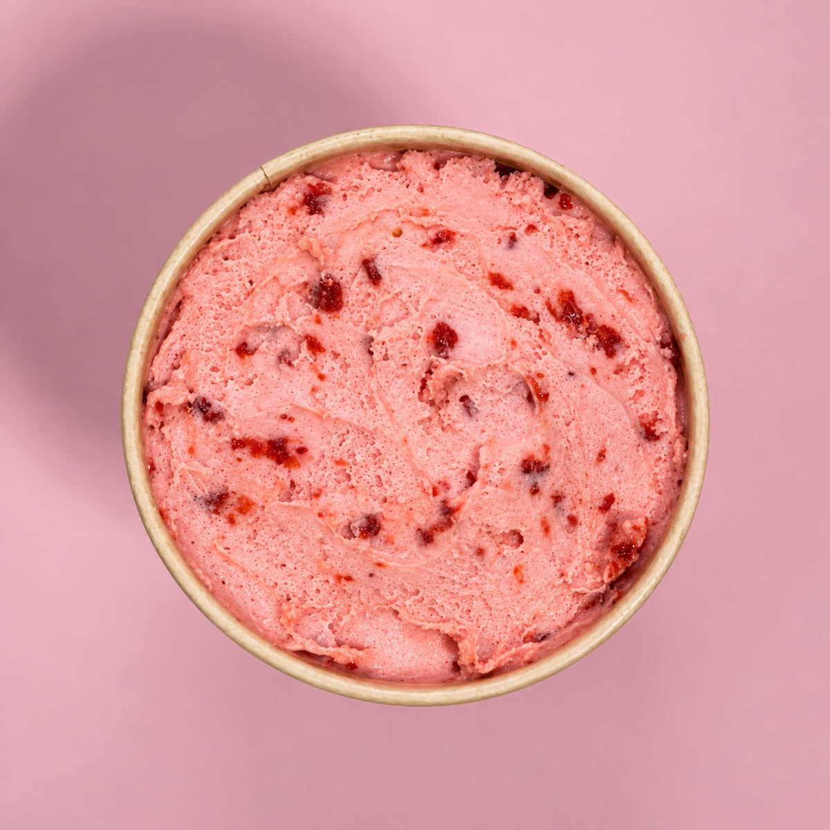 A top-down view of a scoop of pink strawberry ice cream in a light brown paper cup, set against a solid pink background.