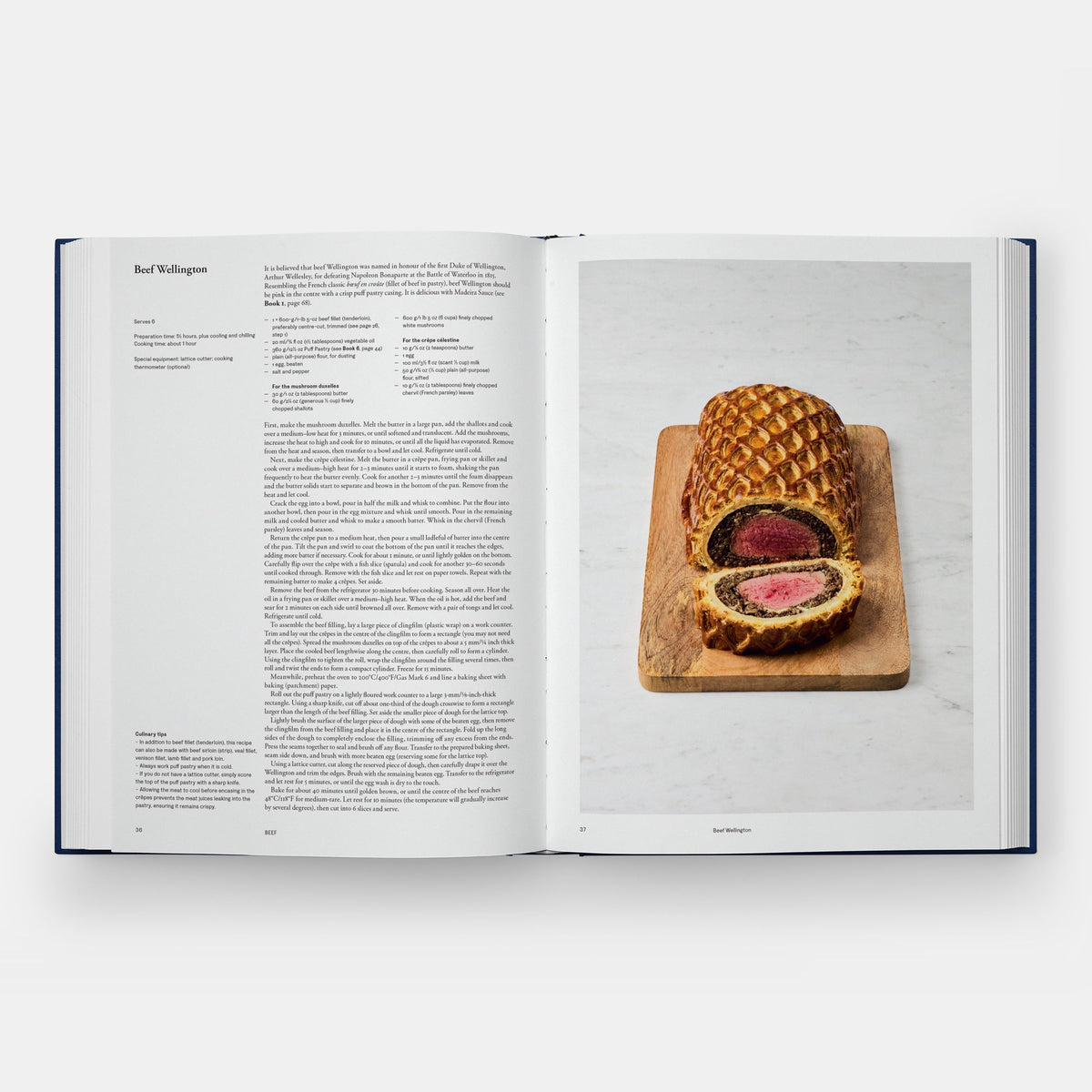 An open cookbook spread showing a recipe page on the left and a plated cut of beef Wellington on a wooden board on the right, photographed against a light gray background.