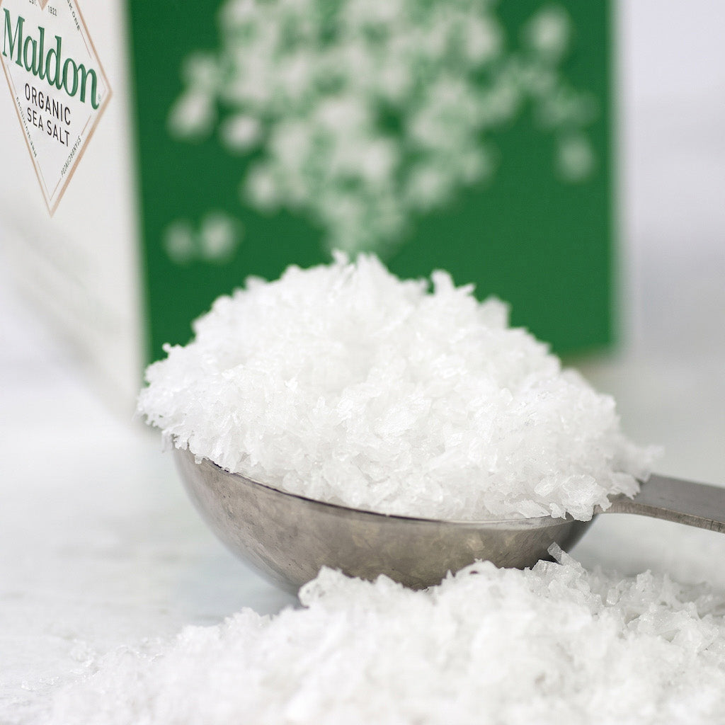 A metal scoop filled with coarse white Maldon sea salt, with a green and white box of Maldon Organic Sea Salt blurred in the background.